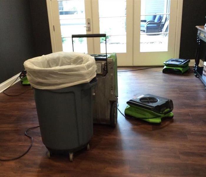 Room with hardwood floor, trash can with white bag.  1 Dehumidifier and 3 air movers on the floor to dry out the room.