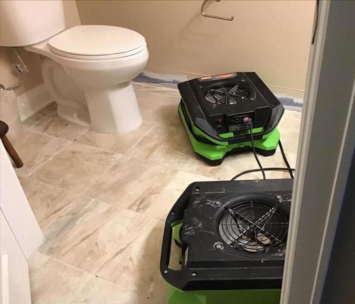 Bathroom square tile, white toilet, baseboard has been removed and two SERVPRO air movers are on the floor.