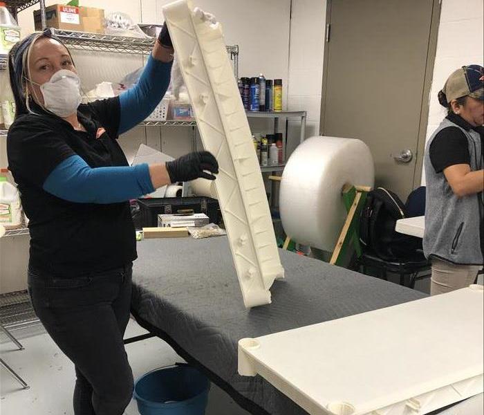 Woman wearing a mask, black pants, black and blue shirt.  She is using a rag to clean a white plastic shelf.
