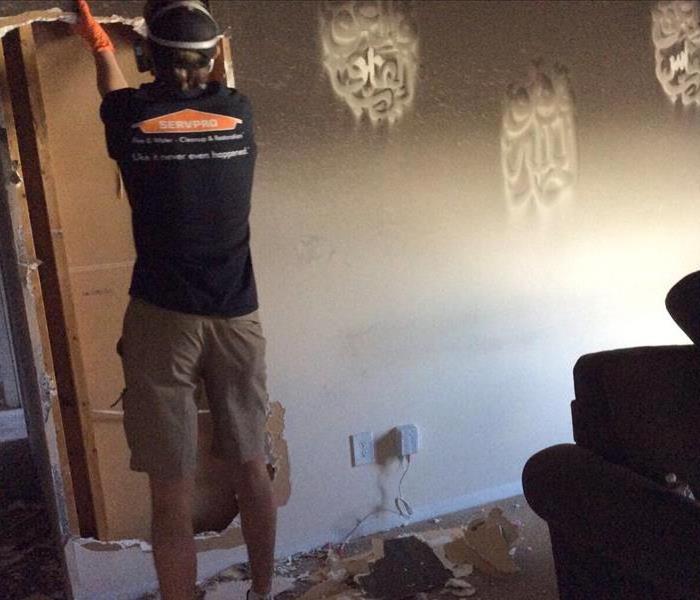Man wearing a black shirt and khaki shorts with a hat and respirator mask pulling down drywall in a room after fire