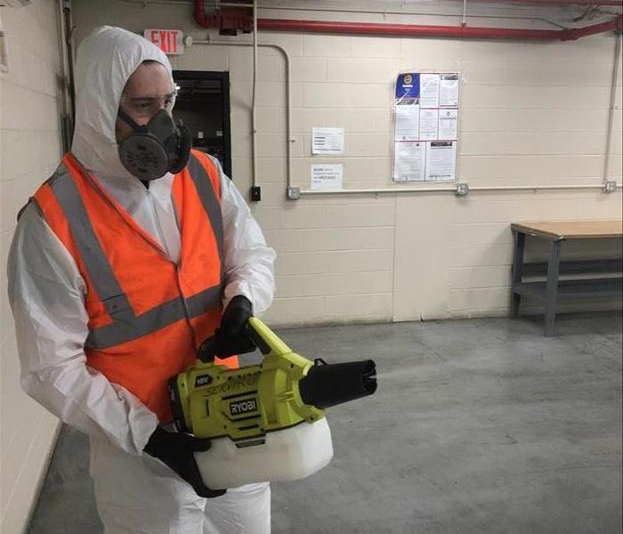 Man wearing a personal protective white suit with orange reflective vest using a commercial fogger machine to clean an area
